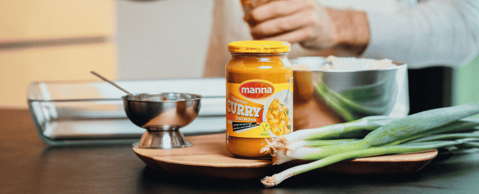 Glazen pot Manna Chinese curry op houten plank, lente-uitjes ernaast en metalen schaaltje op het aanrecht.